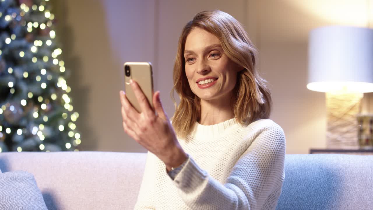 Close Up Portrait Of Happy Female Sitting In Decorated Room Near Christmas Tree With Twinkle Lights And Talking On Video Chat Online On Smartphone On Xmas Eve
