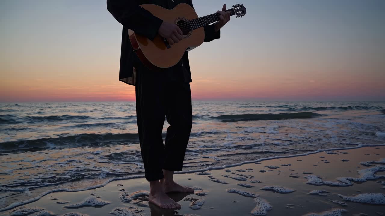 En video i låg vinkel fångar en person som spelar gitarr på en rofylld strand i solnedgången, vilket skapar en lugn blandning