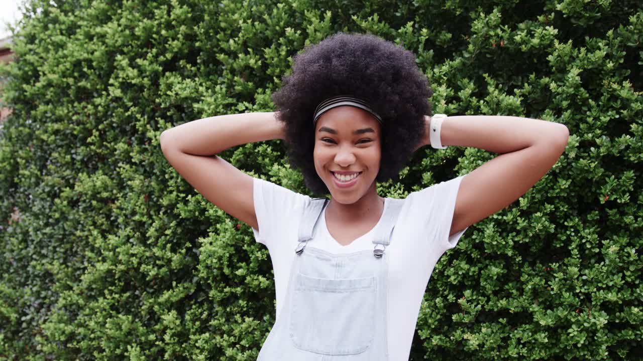 African American woman posing for camera against green hedge in denim overalls, shifting laughing