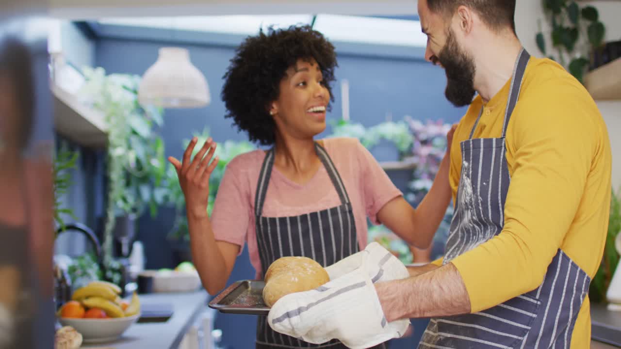 video de una feliz pareja diversa en delantales horneando en la cocina, mirando el pan cocido, con espacio para copiar