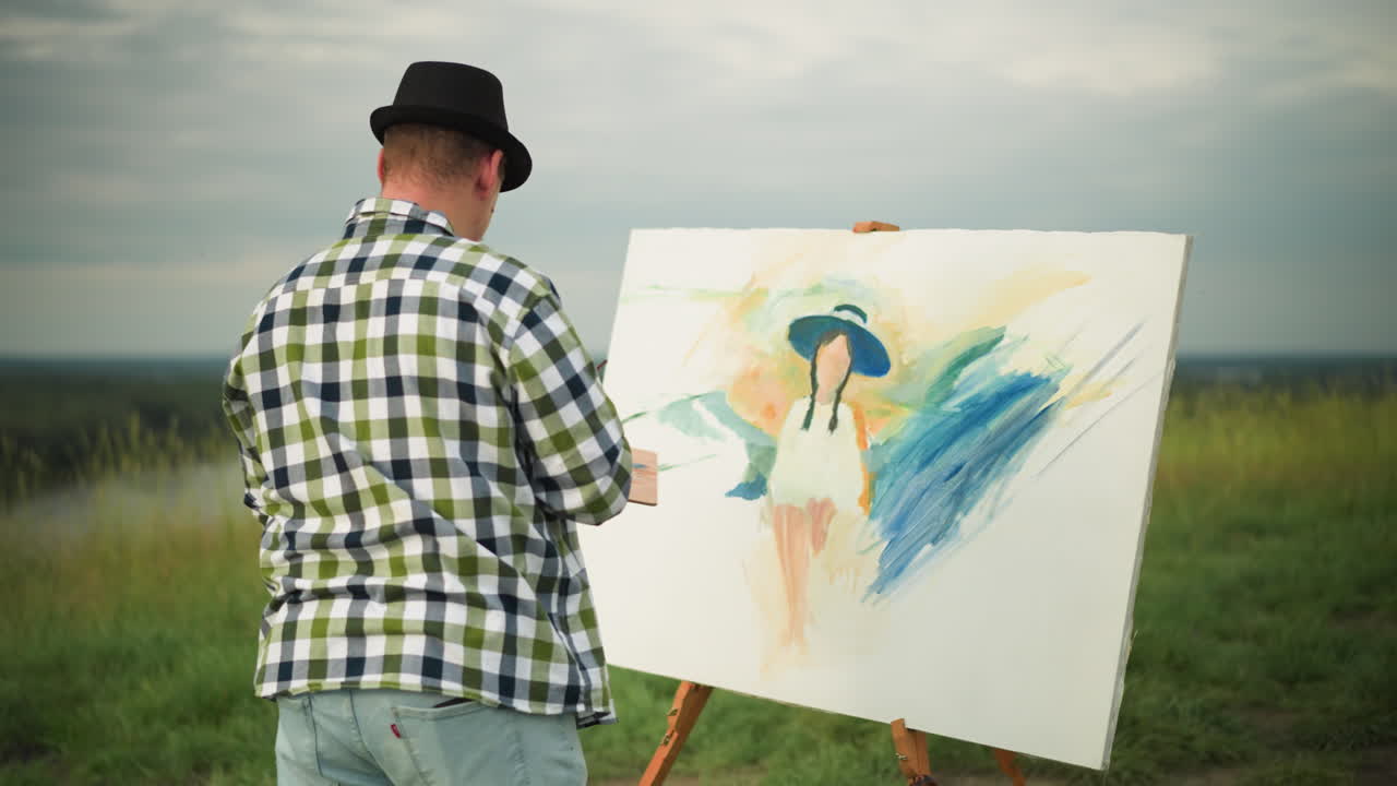 검은 모자와 체크색 셔츠를 입은 예술가가 평화로운 잔디에서 크고 다채로운 캔버스를 칠하는 동안 팔레트를 들고있는 뒷면.
