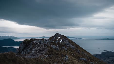 small person standing with arms up in the air on top of a large mountain peak, watching over a beautiful ocean with a grey sky in norway