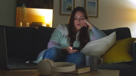 woman sitting on sofa at home at night streaming or watching movie or show on laptop and scrolling internet on phone 5