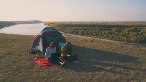 women-with-tablet-and-laptop-sit-near-blue-tent-on-hill