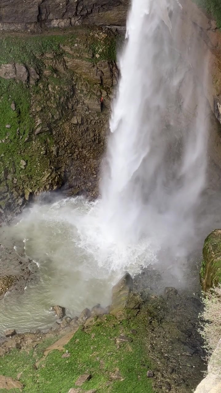 Una potente cascata Tobot scende da una scogliera rocciosa in una pozza sottostante, circondata da erba verde e rocce muschiose.