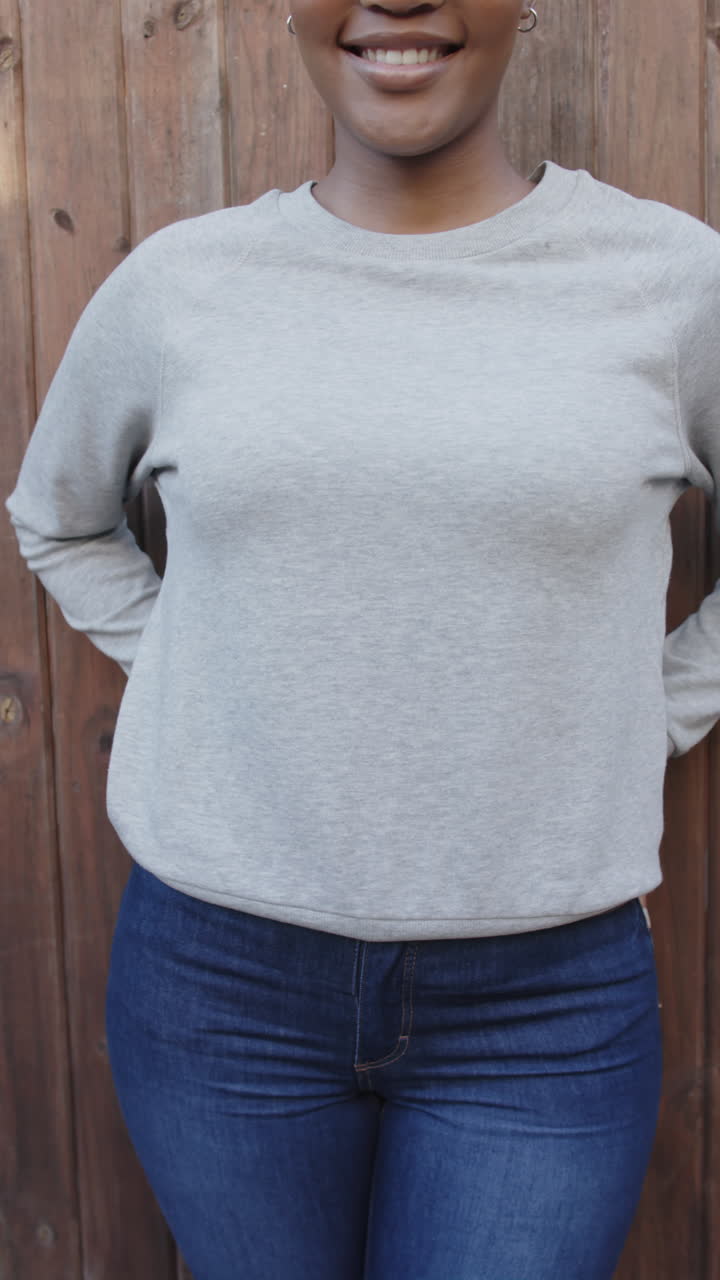 Vertical video of midsection of african american woman in grey sweatshirt against wooden fence