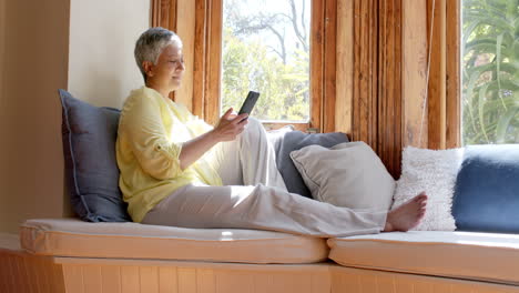 Happy-senior-biracial-woman-sitting-on-couch-and-using-smartphone-at-home,-slow-motion