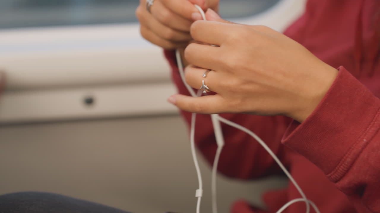 Person carefully untangles headphones. Meticulous individual slowly unties knotted white earphones on lap. Careful person methodically untangles tangled white earbuds resting gently on their lap
