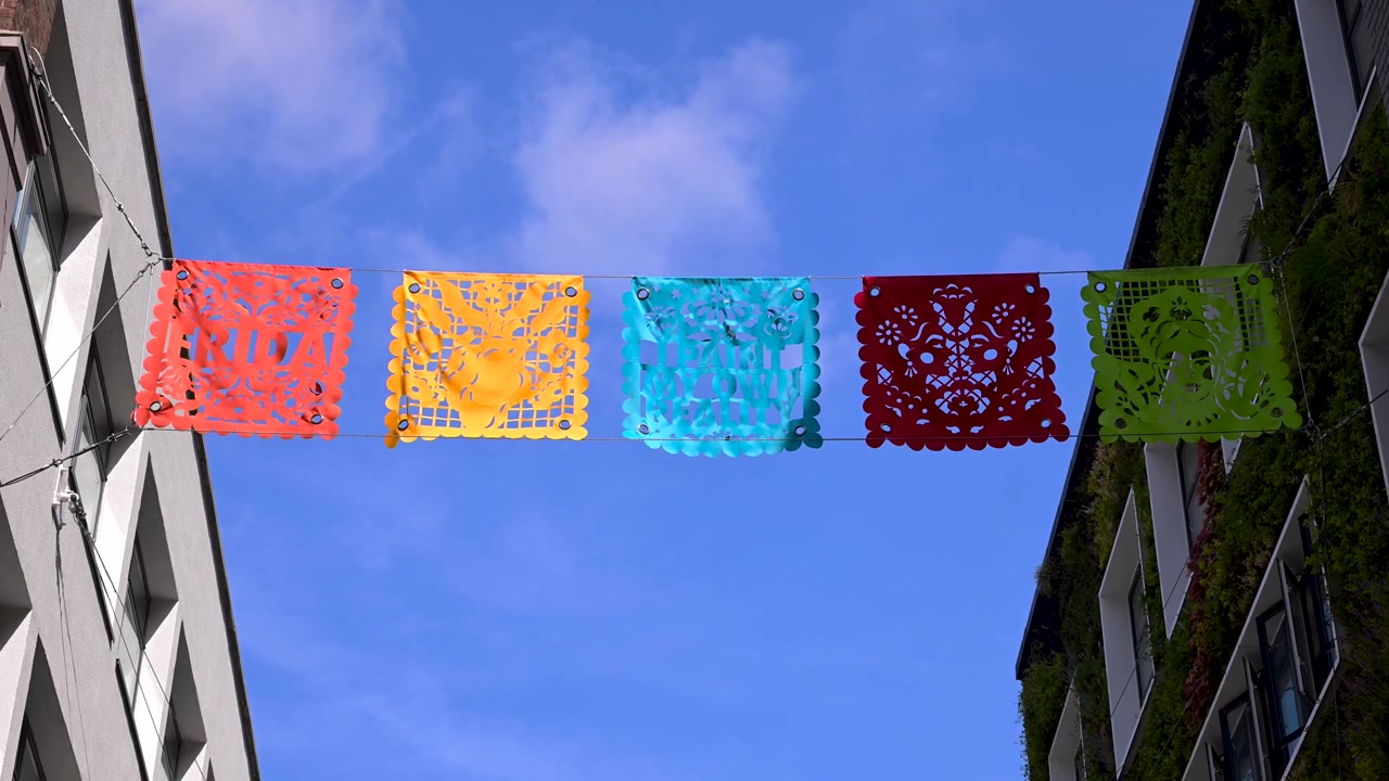 Bandeiras vibrantes de papel picado em vermelho, amarelo, azul, verde-água e verde penduradas entre edifícios urbanos contra um céu azul