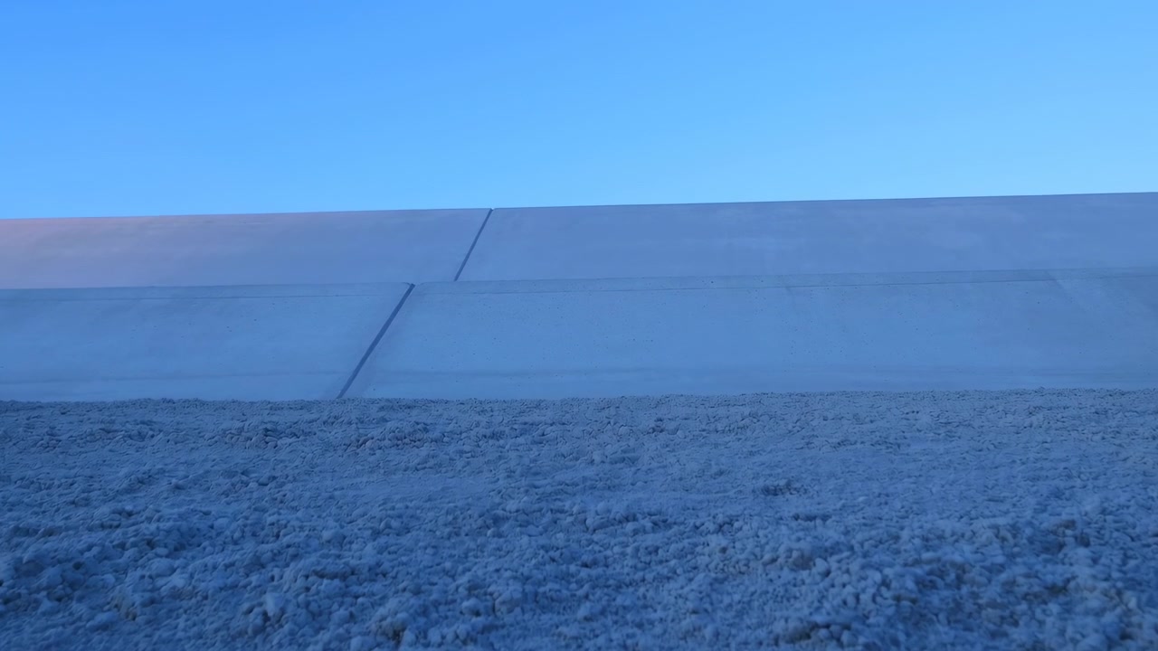 Rough snow blankets foreground beneath long pale concrete barrier crossing horizon, while clear blue sky fills upper view with crisp cold minimal winter mood, with winter, snow, frost, ice detail