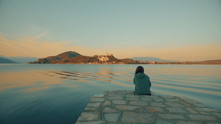 Vista di spalle di una donna con felpa verde con cappuccio seduta sul bordo di un pontile che guarda il Castello di Angera affacciato sul Lago Maggiore in Italia.