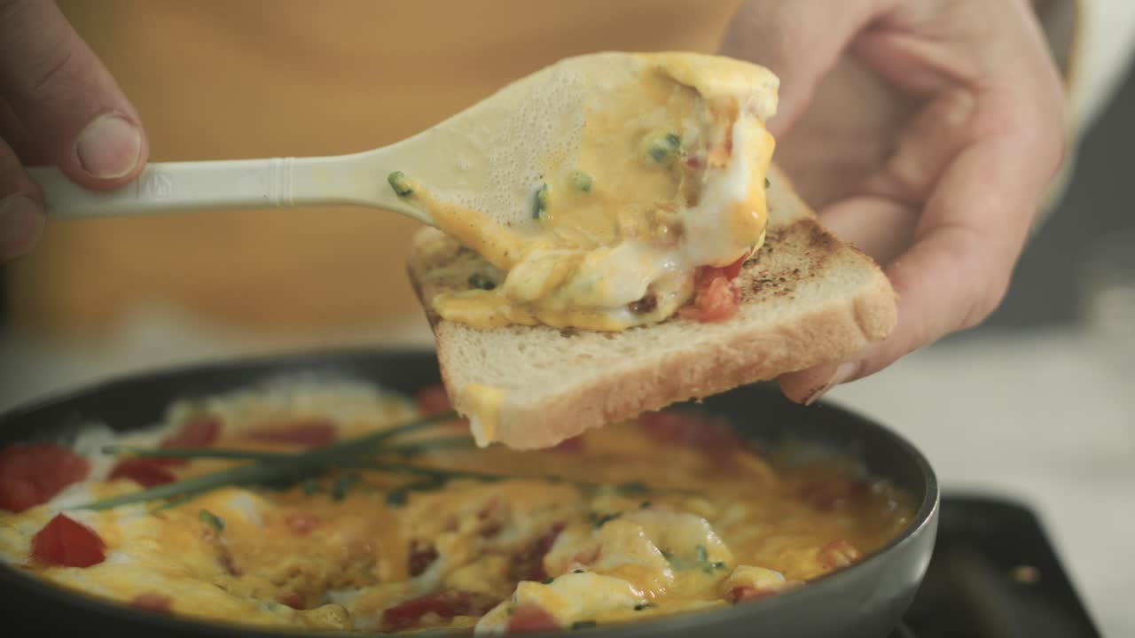 셰프가 갓 요리한 토마토와 골파 조각을 곁들인 비건 스크램블 에그를 나무 숟가락으로 토스트에 올려 놓습니다.
