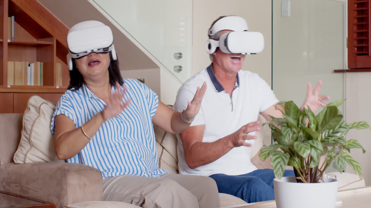 Wearing VR headset, senior diverse couple laughing together on couch at home