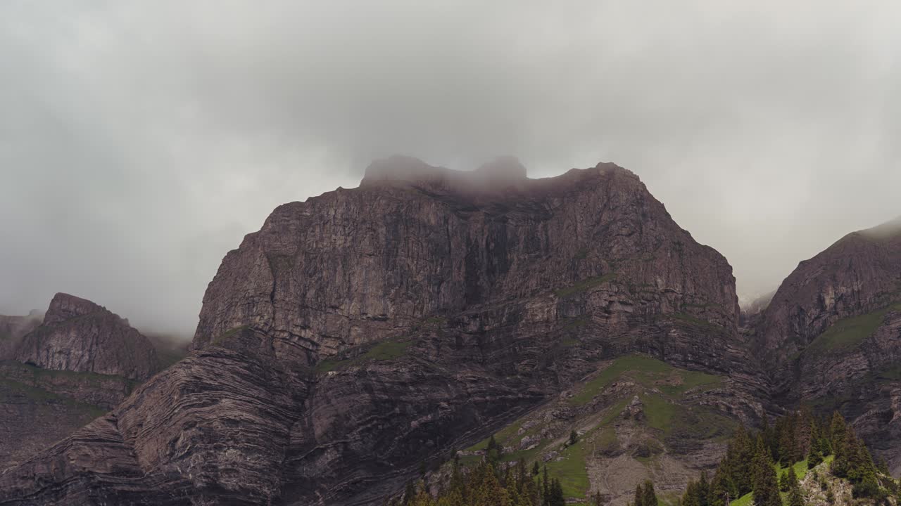 In this timelapse, towering rocky peaks of the Bernese Alps, are partially hidden by low-hanging clouds, with vibrant green alpine meadows adding color to the dramatic, rugged landscape.