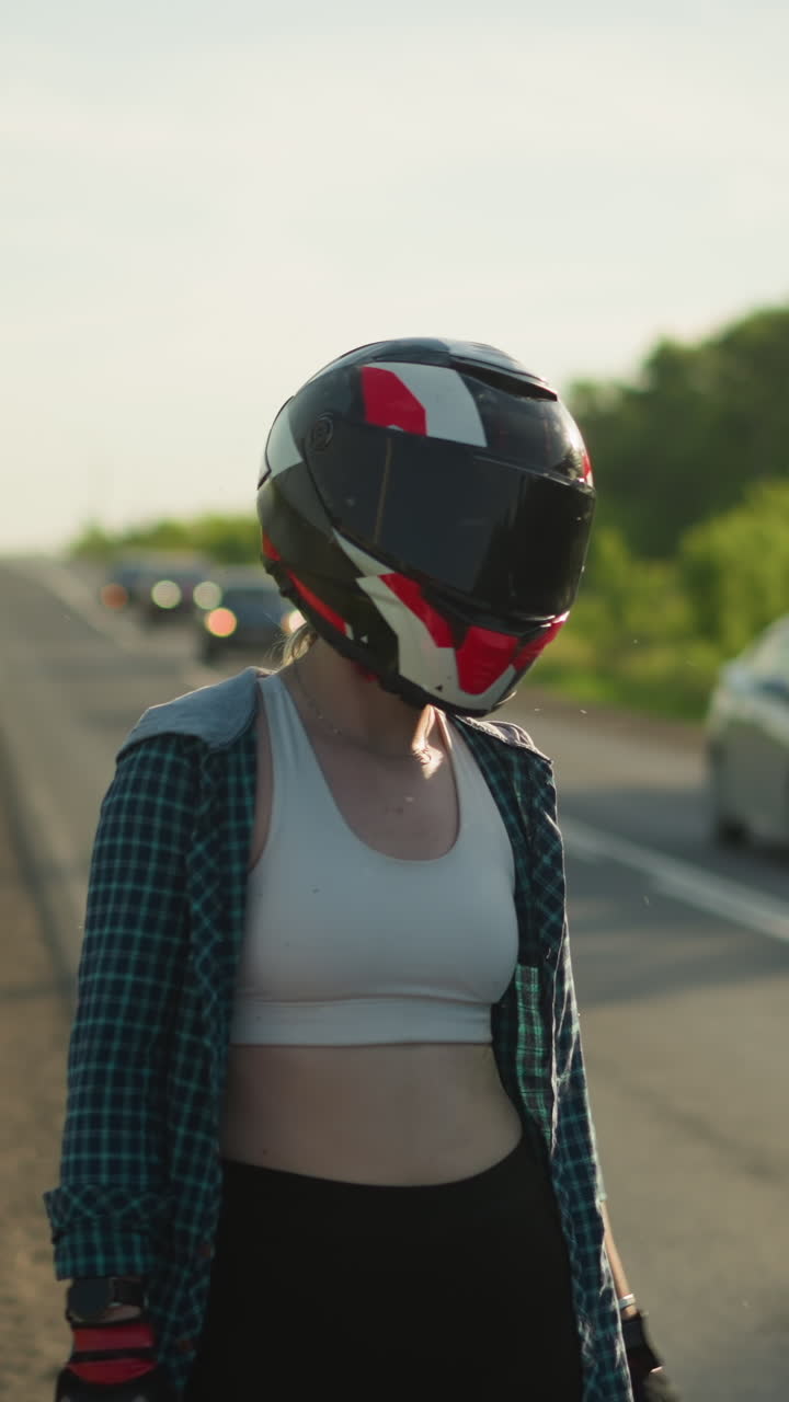 una mujer con un casco de motocicleta está de pie en la carretera, dando un pulgar hacia arriba mientras los coches pasan en la dirección opuesta, está vestida con una camisa a cuadros y una camiseta cortada