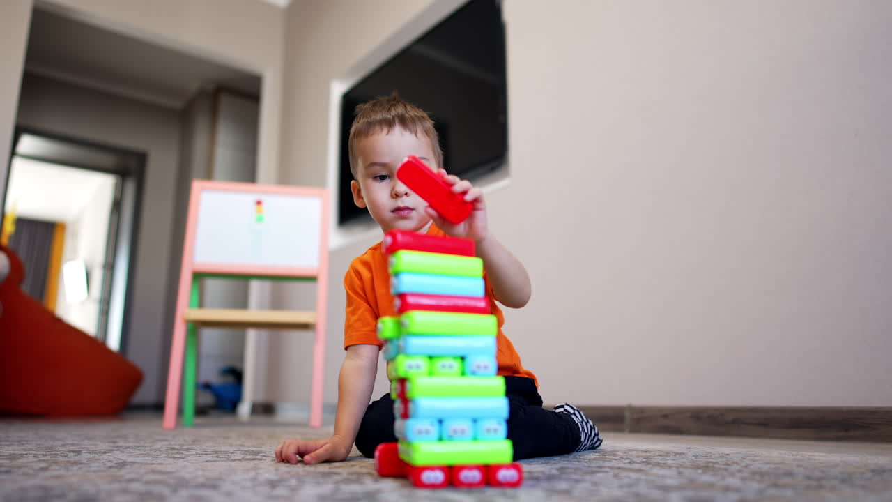 Concentrated Caucasian toddler sits on the floor building a tower. Happy peaceful kid puts blocks on a pile. Low angle view.