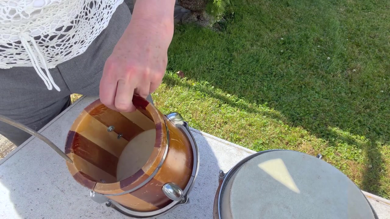 Two handcrafted hand drums with natural animal-hide drumheads rest on a table outdoors. A woman carefully cleans the smaller drum with compressed air, Close-up static shot