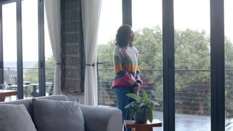 thoughtful biracial woman looking through window at home, slow motion, copy space