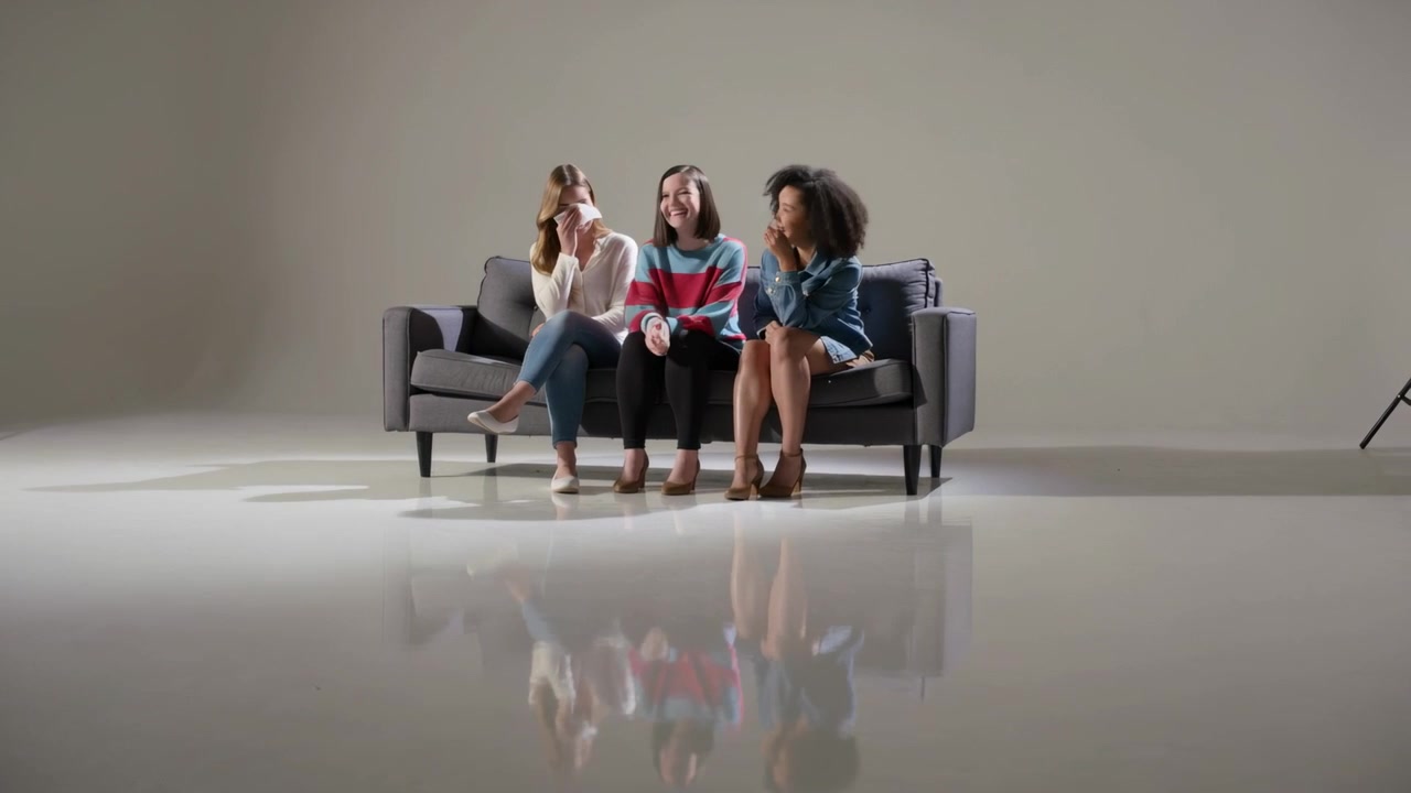 Tre donne su un divano da studio, che reagiscono; quella al centro con una maglietta a righe mostra il telefono, quella a sinistra sorseggia una tazza.