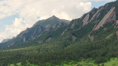 Close-up-aerial-pan-right-of-warm-sun-hitting-Boulder-Colorado-Flatiron-mountains-above-Chautauqua-Park-with-full-green-pine-trees-and-blue-skies-with-clouds-on-a-beautiful-summer-day-for-hiking