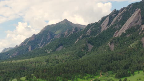 close up aerial pan right of warm sun hitting boulder colorado flatiron mountains above chautauqua park with full green pine trees and blue skies with clouds on a beautiful summer day for hiking