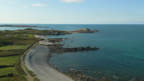 vuelo de aviones no tripulados sobre los páramos y la costa hacia fort doyle, guernsey, calma, aguas cristalinas, playas de guijarros y formaciones rocosas y una torre martello con vistas al mar en un día soleado.