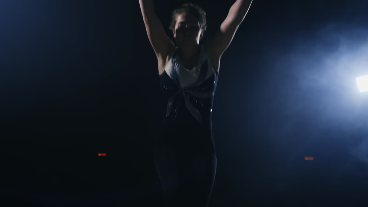 A young female gymnast runs up looking at the camera and performs a forward flip and screw in the air in slow motion in the gym on the carpet in the smoke. the Steadicam in motion and backlight