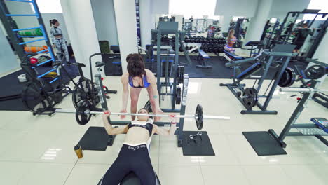 determined athlete lying on the bench and lifting barbell. sportswoman does inclined dumbbell presses and sexy female instructor helps her.