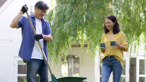 Reifer-Asiatischer-Mann-Harkt-Und-Räumt-Blätter-Im-Garten-Auf,-Während-Seine-Frau-Ihm-Ein-Heißes-Getränk-In-Einer-Tasse-Bringt-–-Aufnahme-In-Zeitlupe