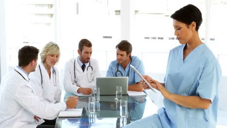 Thoughtful-surgeon-using-her-tablet-with-staff-talking-behind-her