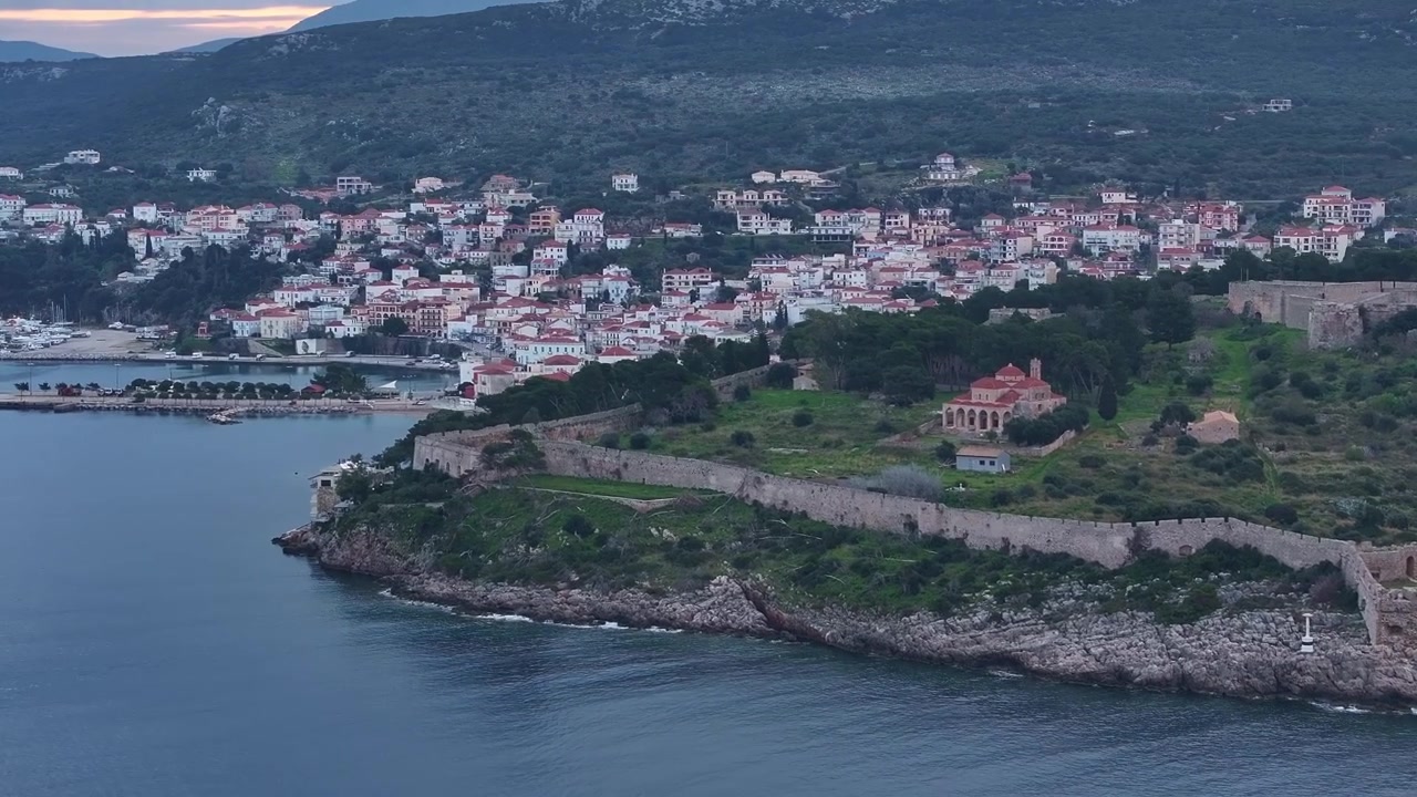 Panoramica aerea con drone lungo le antiche mura in pietra della fortezza di Niokastro, che rivela la Chiesa della Trasfigurazione, il porto costiero e la città illuminata di Pylos al crepuscolo in Grecia.