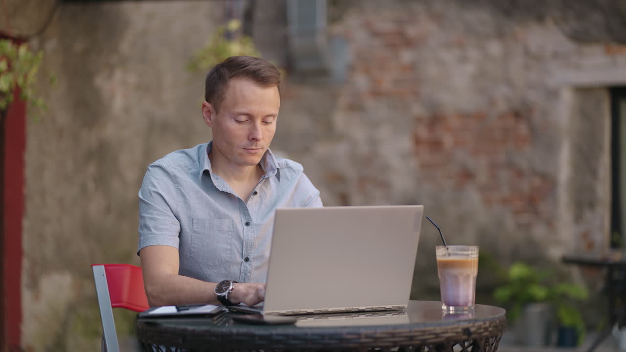 A young man in a shirt is sitting at a table with a laptop and typing on the keyboard. A student can study remotely. A businessman conducts his business remotely