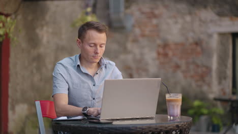 Un-Joven-Con-Camisa-Está-Sentado-En-Una-Mesa-Con-Una-Computadora-Portátil-Y-Escribiendo-En-El-Teclado.-Un-Estudiante-Puede-Estudiar-De-Forma-Remota.-Un-Empresario-Realiza-Su-Negocio-De-Forma-Remota