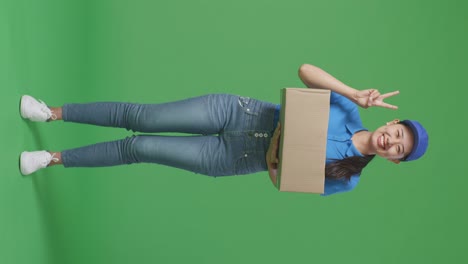 full body of asian female courier in blue uniform showing gesture peace and smiling while delivering a carton on green screen background in the studio