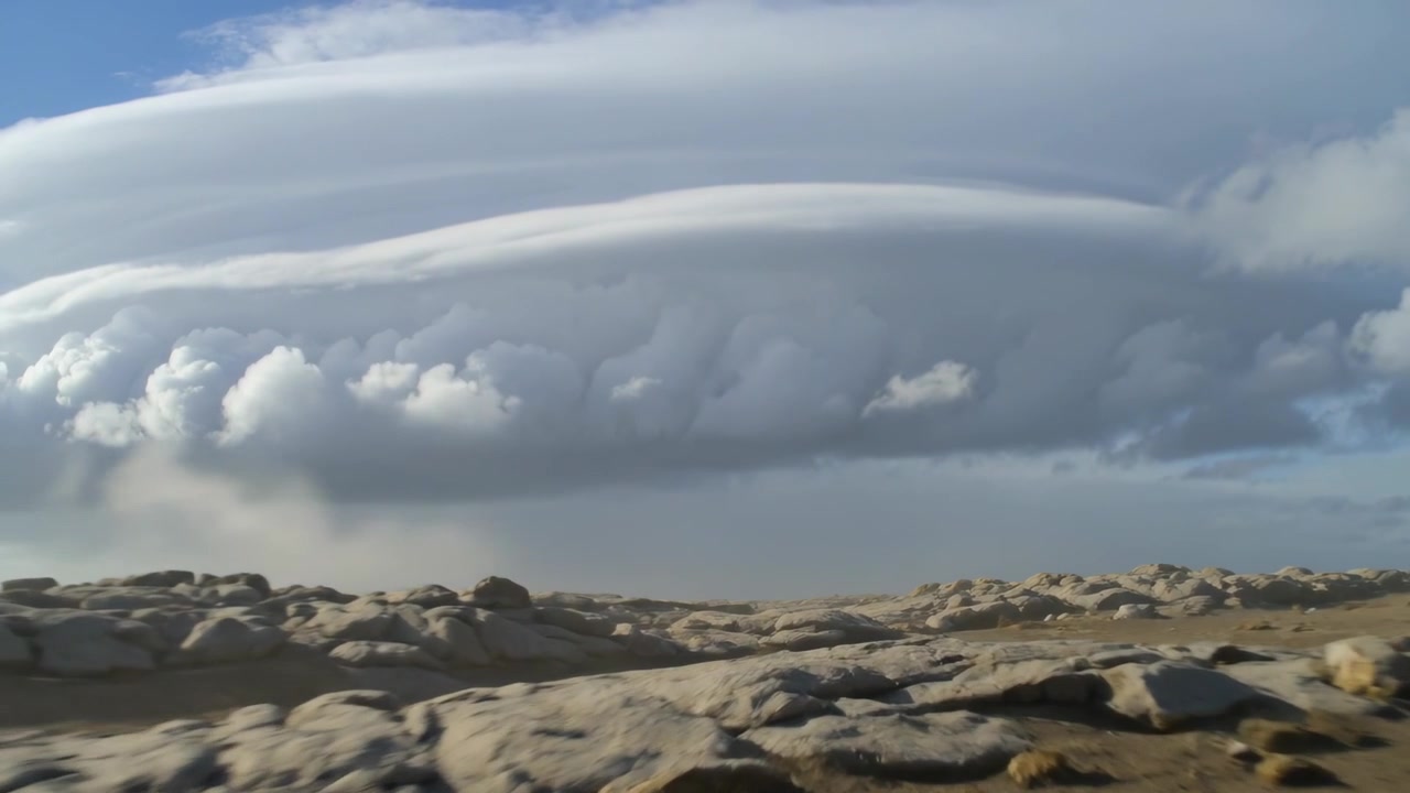 Panorerende kamera afslører en stor, lagdelt linseformet sky over en stenet kystslette med svag virga.