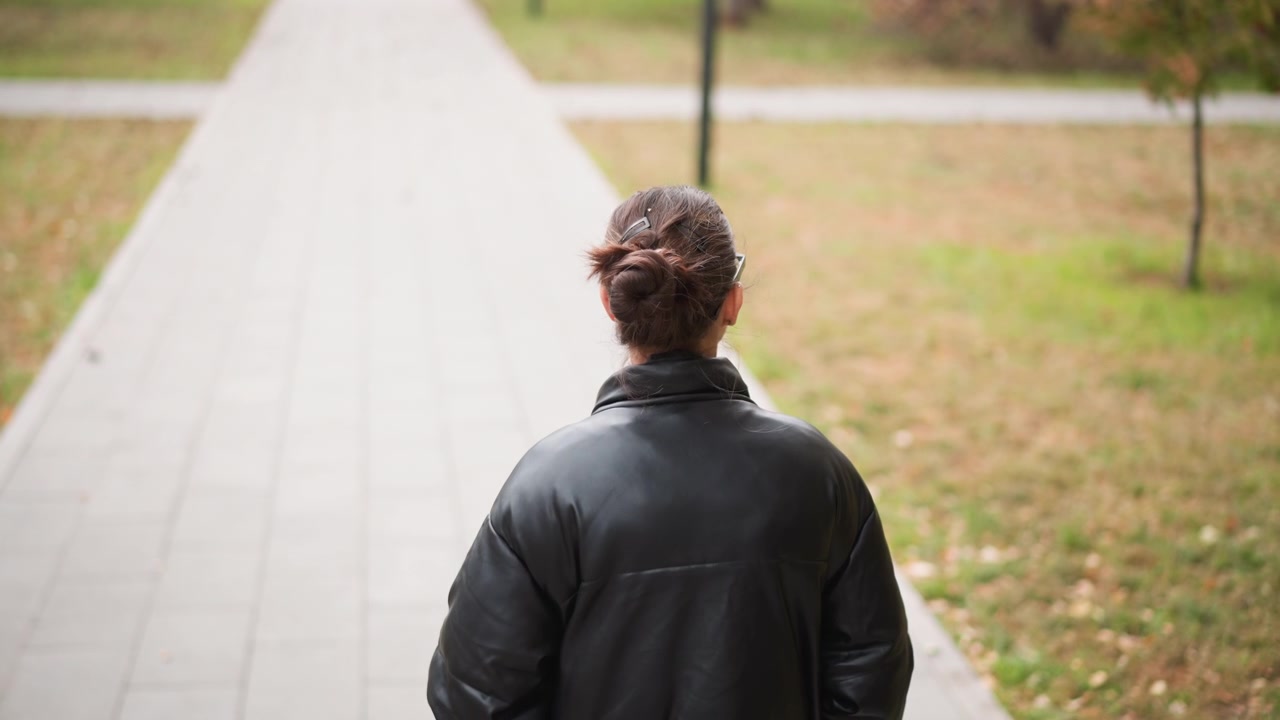 Close up of woman with dark hair in bun wearing glasses and black leather jacket walking steadily away on paved path. background features grassy areas scattered fallen leaves trees