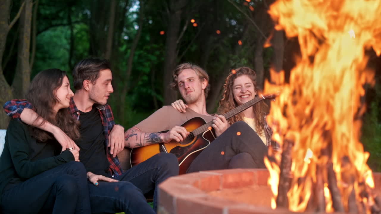 A group of happy young friends near a campfire at glamping, night. Two men and women. Playing guitar. Slow motion