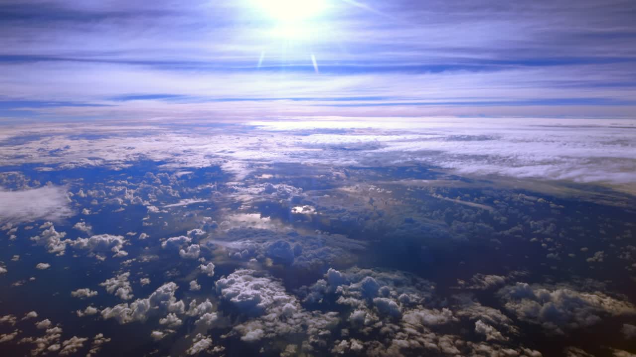 A high altitude aerial view above the clouds on a sunny morning, taken from the cabin of a passenger aircraft. The camera slowly tilt up, looking out the port side window as the plane flies leveled.