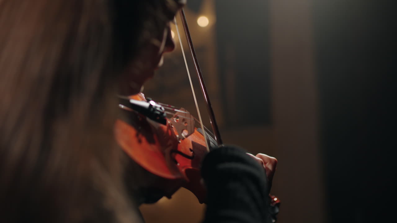 una mujer violinista con ropa negra está tocando música clásica en una sala filarmónica oscura.