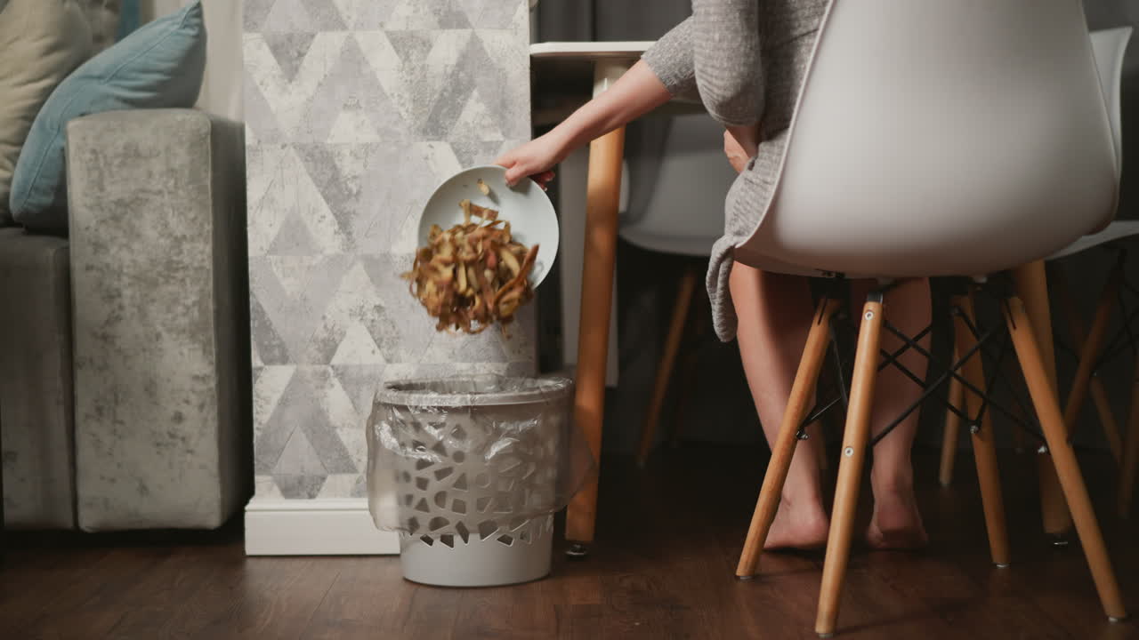 Back view of person sitting on white chair carefully disposing trash into waste bin next to wall with stylish geometric wallpaper, wooden floor and cozy home interior setting in background