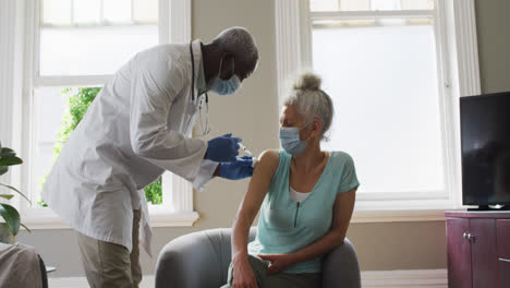 male african american doctor wearing face mask injecting covid-19 vaccine into senior female patient