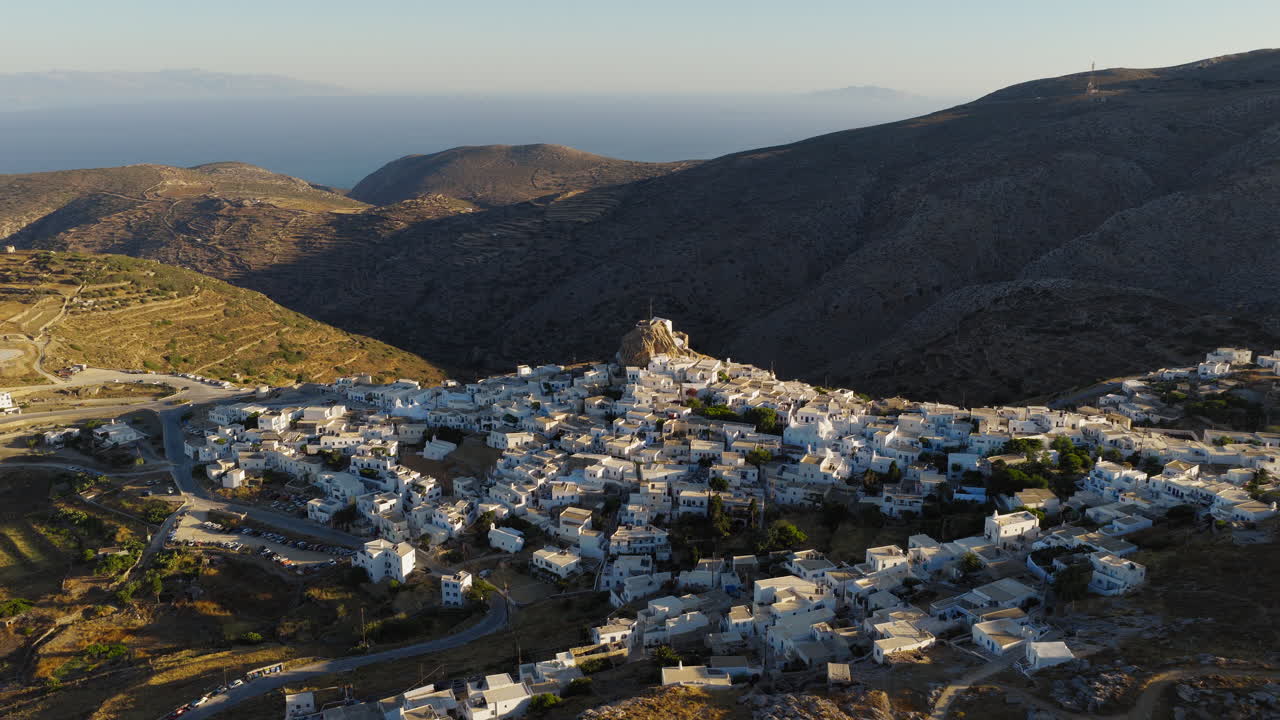 Panoramic drone footage of Amorgos Chora at sunrise, flying toward the historic Amorgos Castle as golden light bathes the Cycladic village