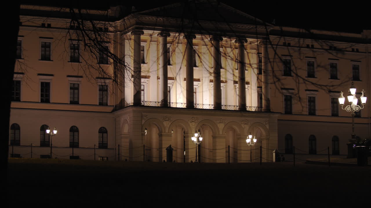 Slow motion close-up to wide 4K shot with parallax motion of dark trees moving in front of illuminated Norwegian Royal Palace on top of Karl Johan street, at night in Oslo Norway