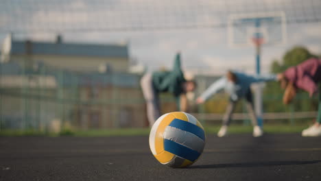 close-up of volleyball resting on outdoor court with blurred view of athletes stretching in background, sunlight casts warm tones on ball, with sports facility and buildings in the background