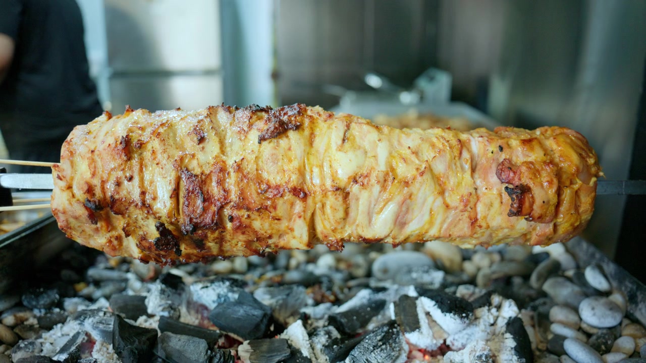 Carne marinata impilata che arrostisce lentamente su uno spiedo verticale su carboni ardenti, preparando popolare cibo da strada.