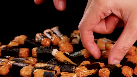 a hand reaches into a pile of roasted rice crackers wrapped in seaweed, picking up a piece. close-up, studio lighting, black background