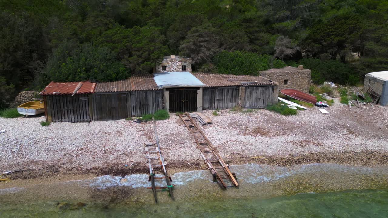 Cala Llentrisca, a secluded beach in Ibiza with Historic boat garage, highlighting the pristine turquoise waters and surrounding verdant landscape. Best aerial view flight static tripod hovering drone