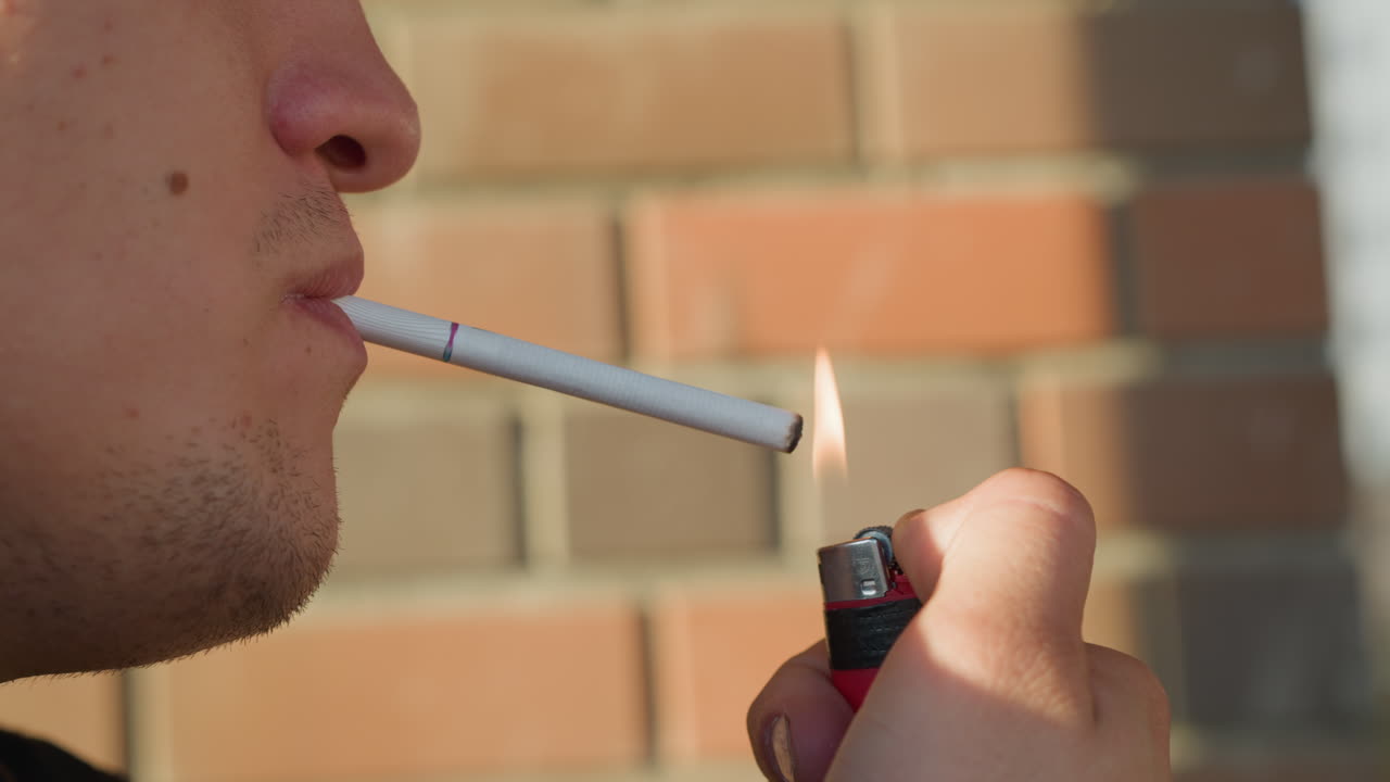 close up of boy holding cigarette in mouth while lighting it with open flame from red lighter against sunlit outdoor brick wall background during daytime