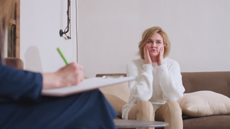 guest seated on sofa talking to counselor while counselor takes note during reflective session, showing professional dialogue and emotional expression in calm therapeutic setting with soft lighting
