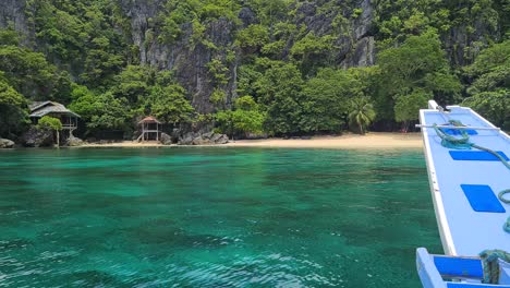 Boat-Arriving-on-Exotic-Tropical-Beach-With-Abandoned-House-Under-Steep-Limestone-Cliffs